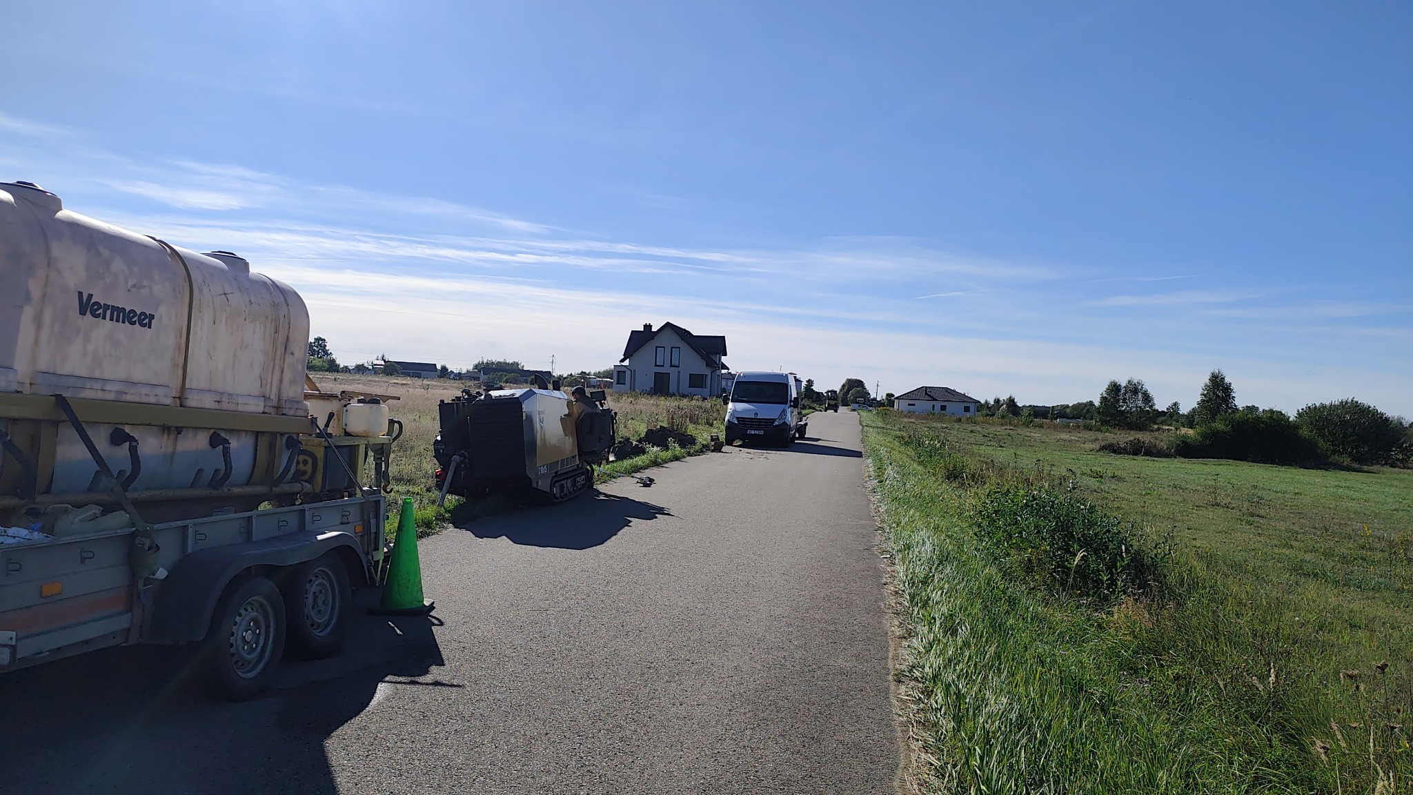 Przewiert sterowany Vermeer na przyczepie, wykop i bus na drodze wiejskiej, błękitne niebo. Sprzęt do instalacji podziemnych w trakcie pracy.