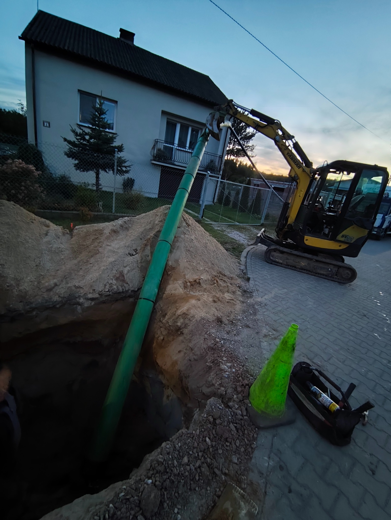 Żółta koparka Yanmar umieszcza zieloną rurę w wykopie na tle domu jednorodzinnego. Widoczny stożek ostrzegawczy i torba narzędziowa.