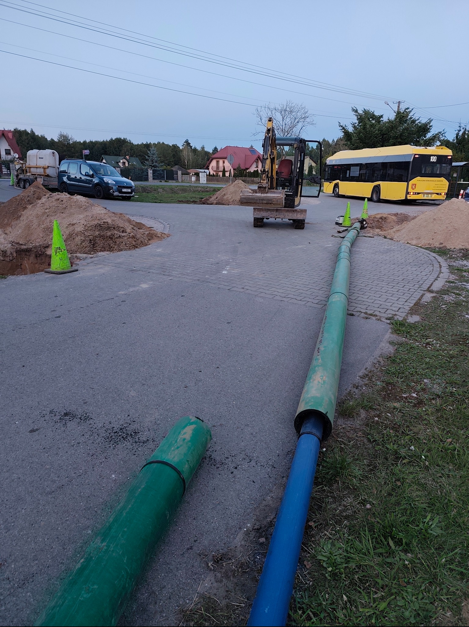 Prace ziemne: wykop, rury wodociągowe (zielone i niebieskie), mała koparka Yanmar, hałdy piachu i żółty autobus w tle. Prace przyłączeniowe w toku.