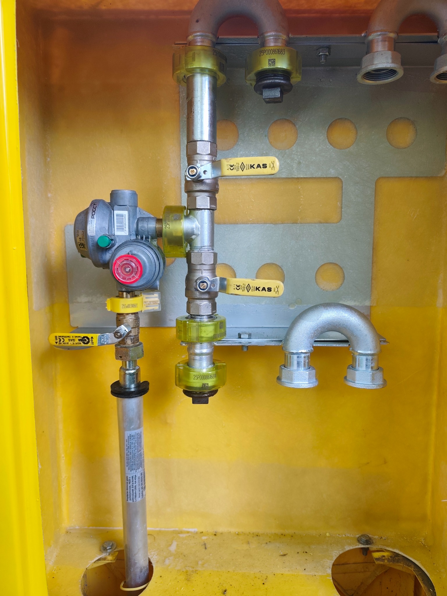 Żółta szafka gazowa z zaworami, reduktorem i rurami. Widoczne elementy instalacji gazowej z oznaczeniami i numerami seryjnymi. Detal przyłącza gazowego wewnątrz skrzynki.