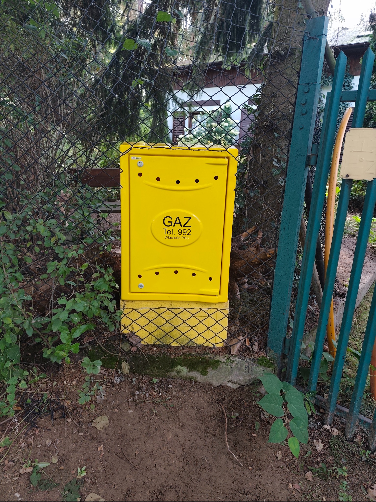 Żółta skrzynka gazowa z napisem 'GAZ' i numerem telefonu, umieszczona przy ogrodzeniu z siatki, na tle zieleni i fragmentu budynku.