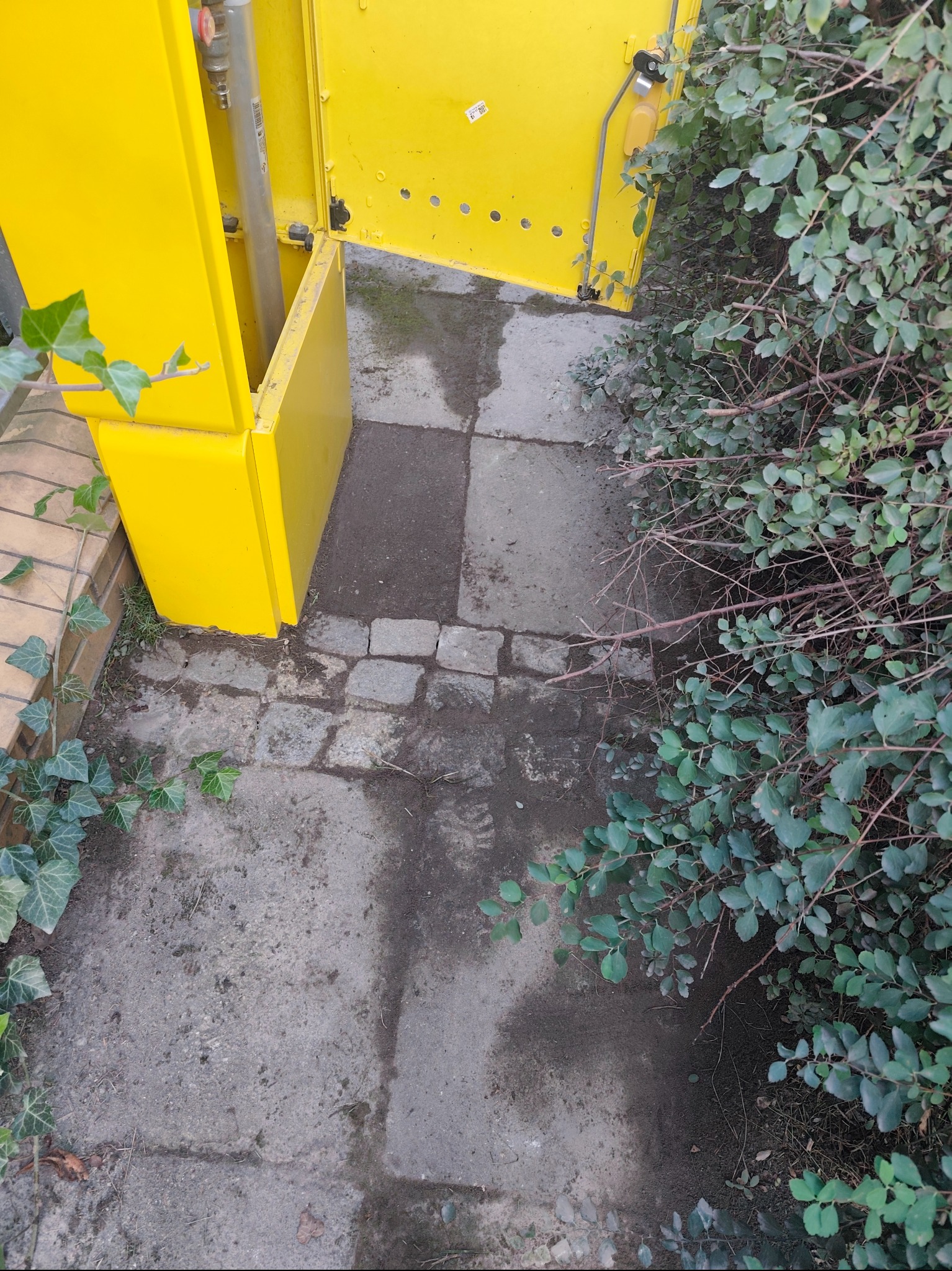 Otwarta, żółta skrzynka gazowa z widoczną rurą, umieszczona na betonowej ścieżce obok zieleni. Kąt ujęcia z góry, oświetlenie naturalne.