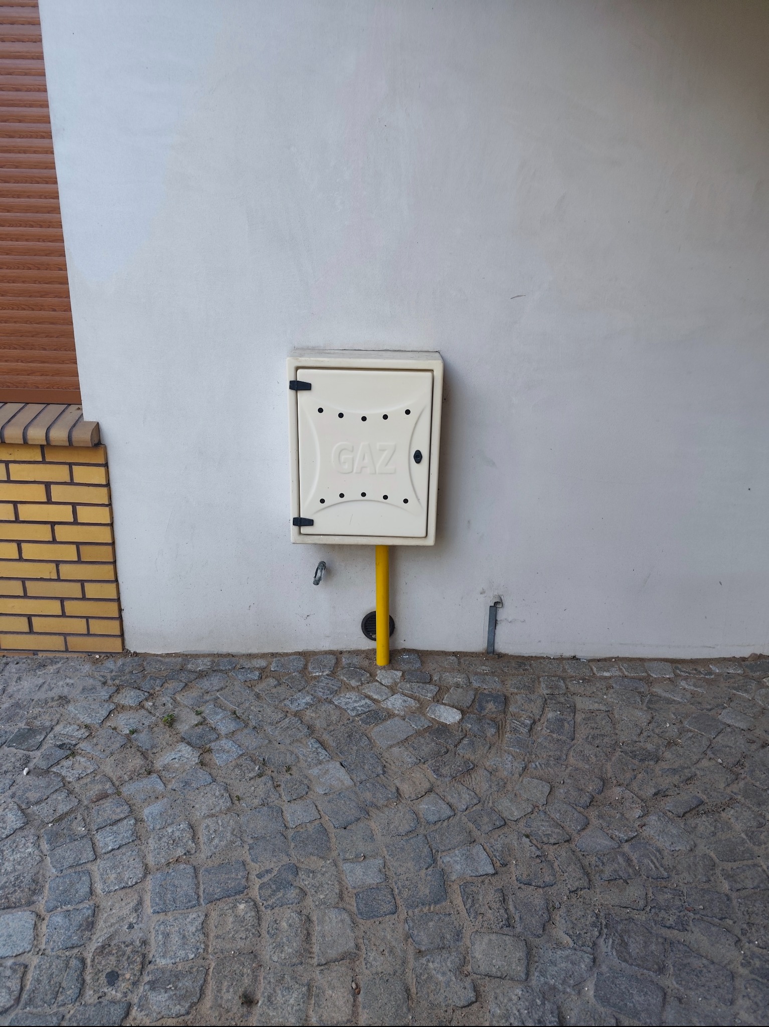 Skrzynka gazowa z napisem 'GAZ' na elewacji budynku, z żółtą rurą. Kostka brukowa na dole kadru. Widoczne elementy instalacji gazowej.