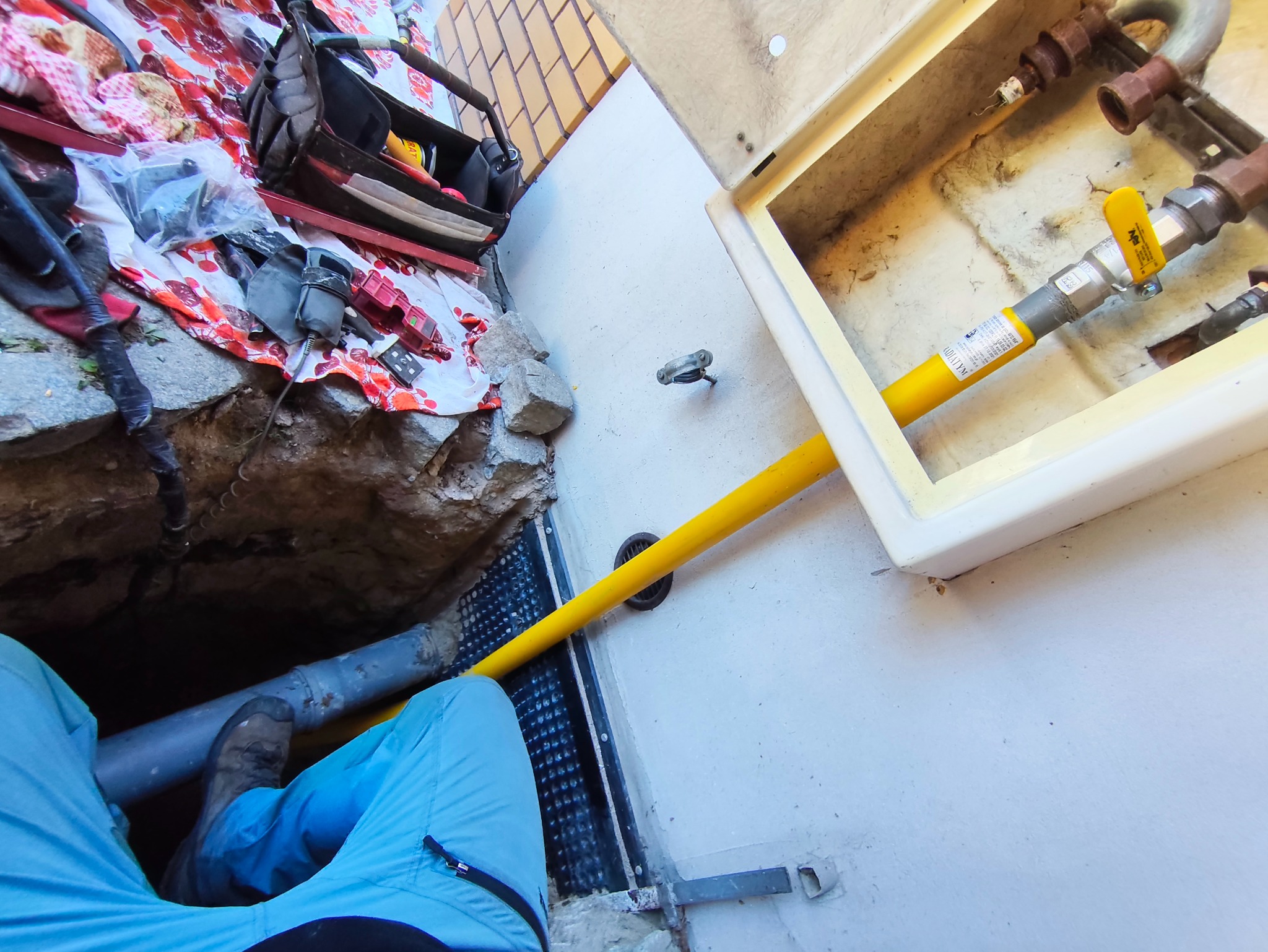 Żółta rura gazowa wchodzi do skrzynki przyłączeniowej na ścianie budynku. Obok narzędzia i wykop. Widok z góry, ukośnie. Prace instalacyjne w toku.