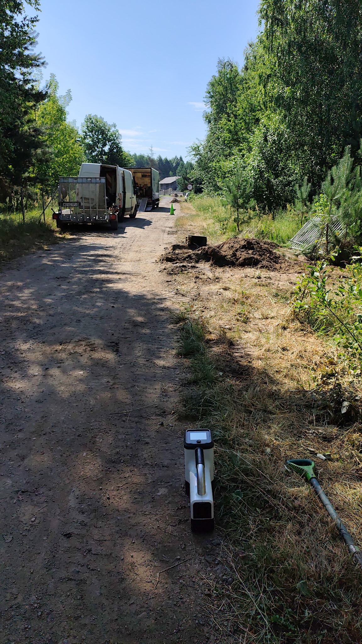 Przenośny analizator gruntu i łopata na ziemistej drodze w lesie, w tle samochody dostawcze i wykopana ziemia. Sprzęt do badań geotechnicznych w terenie.