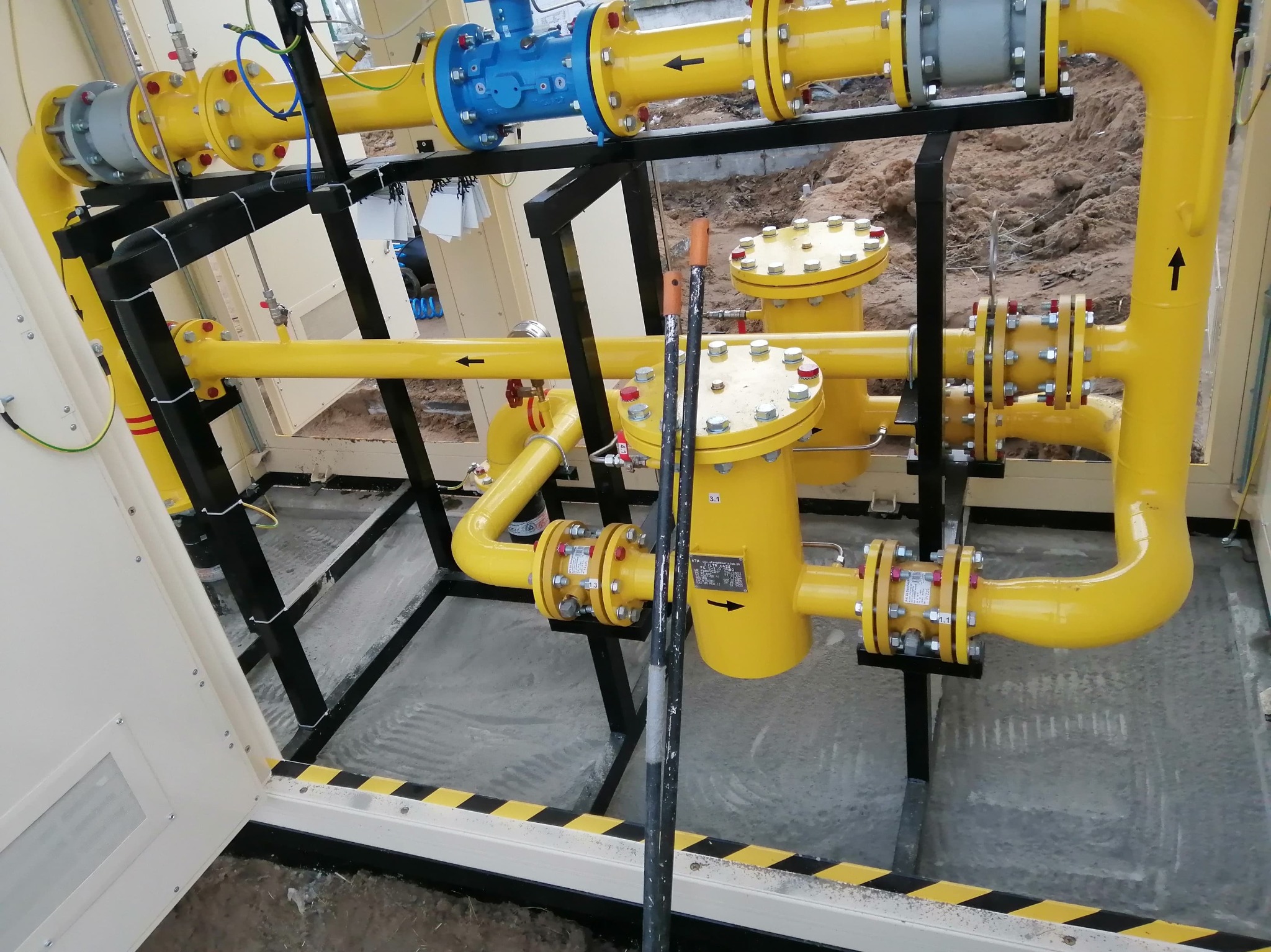 Żółte rury gazowe z zaworami i czarnymi strzałkami, zamontowane na czarnej konstrukcji. Widoczne połączenia śrubowe i fragment podłoża z pasami ostrzegawczymi.