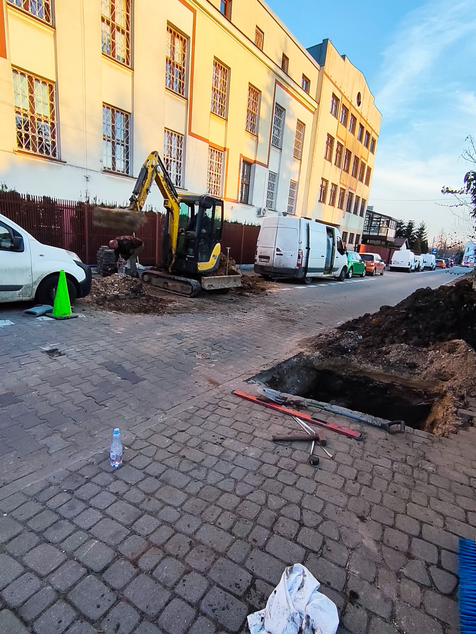 Żółta mini koparka przy wykopie na ulicy z kostki, w tle budynek i zaparkowane samochody. Widoczne narzędzia i akcesoria budowlane.