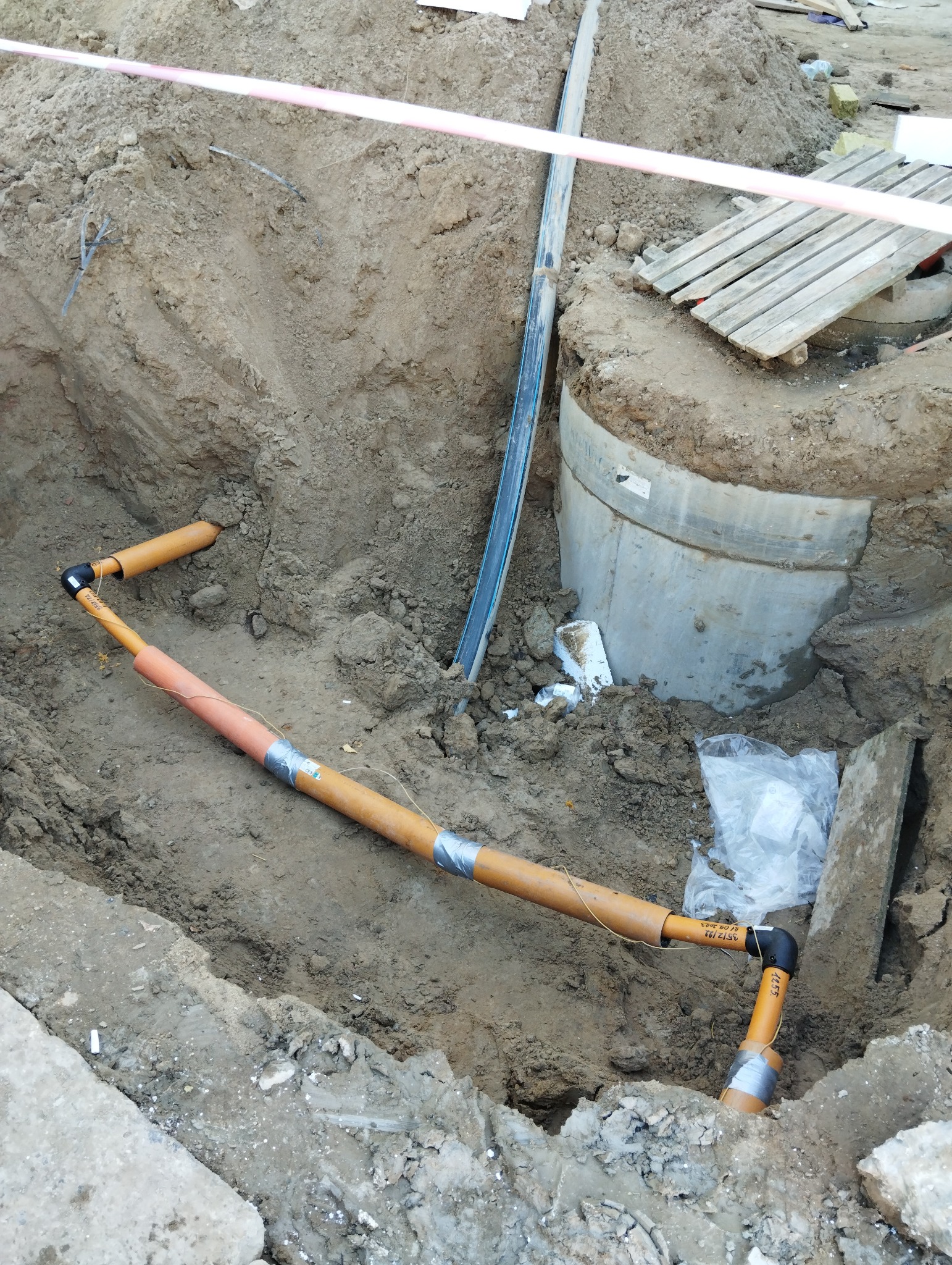 Wykonany fragment instalacji wodnej z rur w wykopie ziemnym, zabezpieczonych taśmą. Widoczny betonowy element studni i taśma ostrzegawcza.