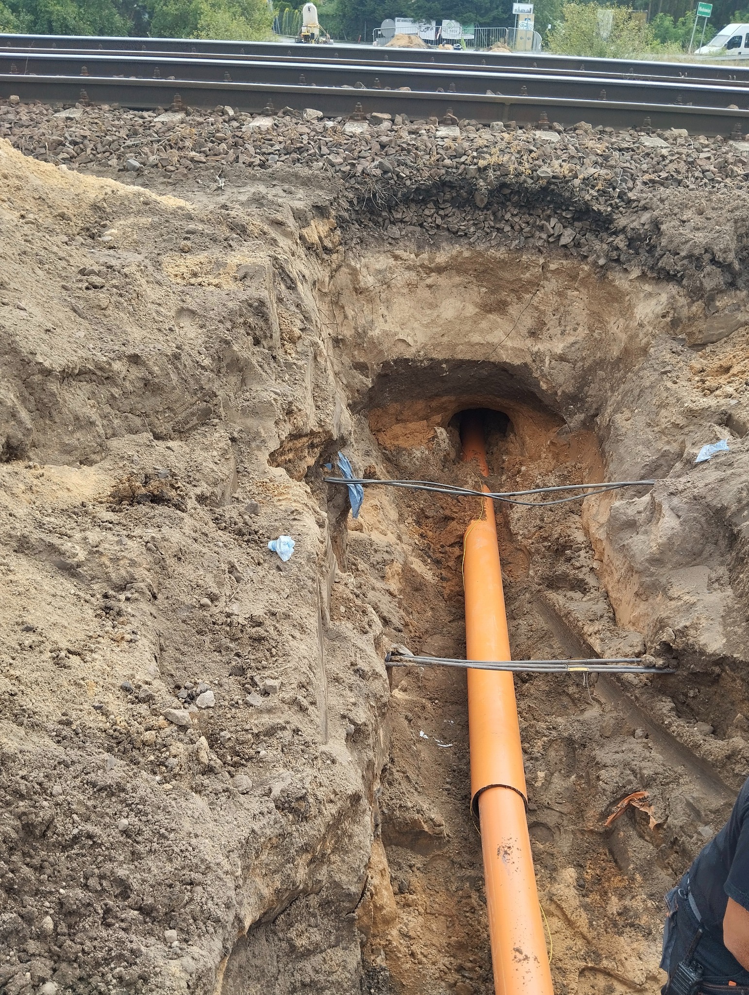 Pomarańczowa rura w wykopie pod torami kolejowymi, zabezpieczona stalowymi linkami. Widoczny tunel pod nasypem.