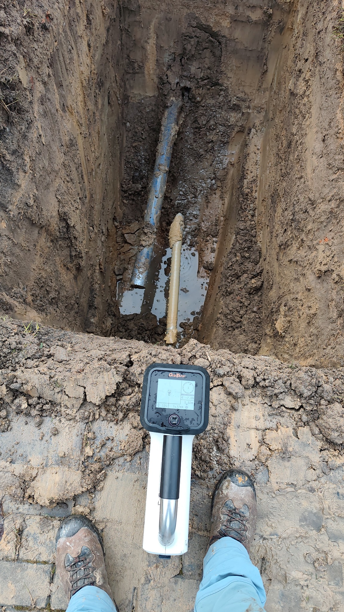 Wykop z rurami i wodą, widoczny detektor podziemnych instalacji. Ujęcie z góry, z butami osoby wykonującej pomiar. Błoto na ziemi i urządzeniu.
