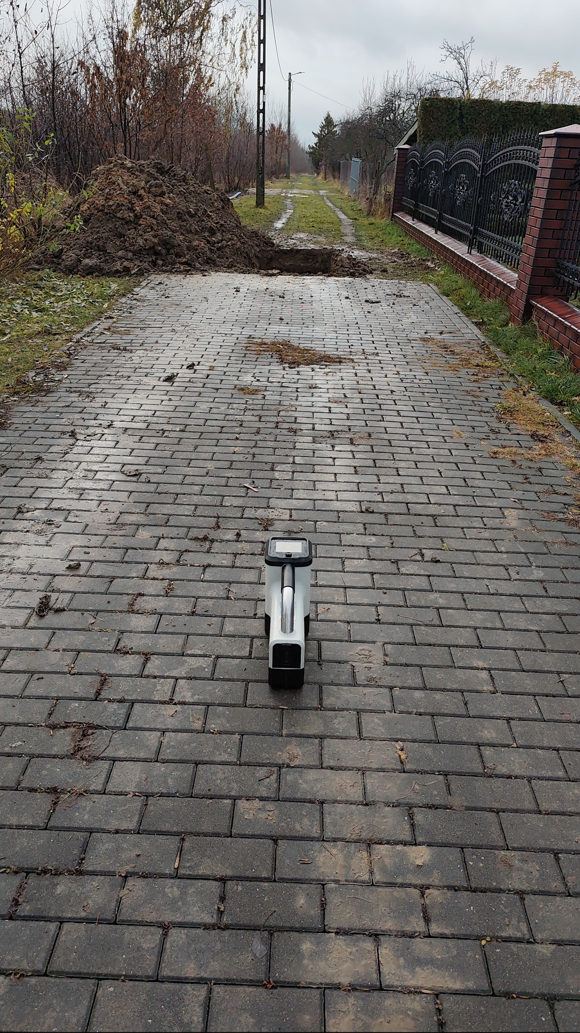 Wykop w brukowanej drodze z hałdą ziemi i urządzeniem pomiarowym. Widok w perspektywie, pochmurne niebo, jesienna sceneria.