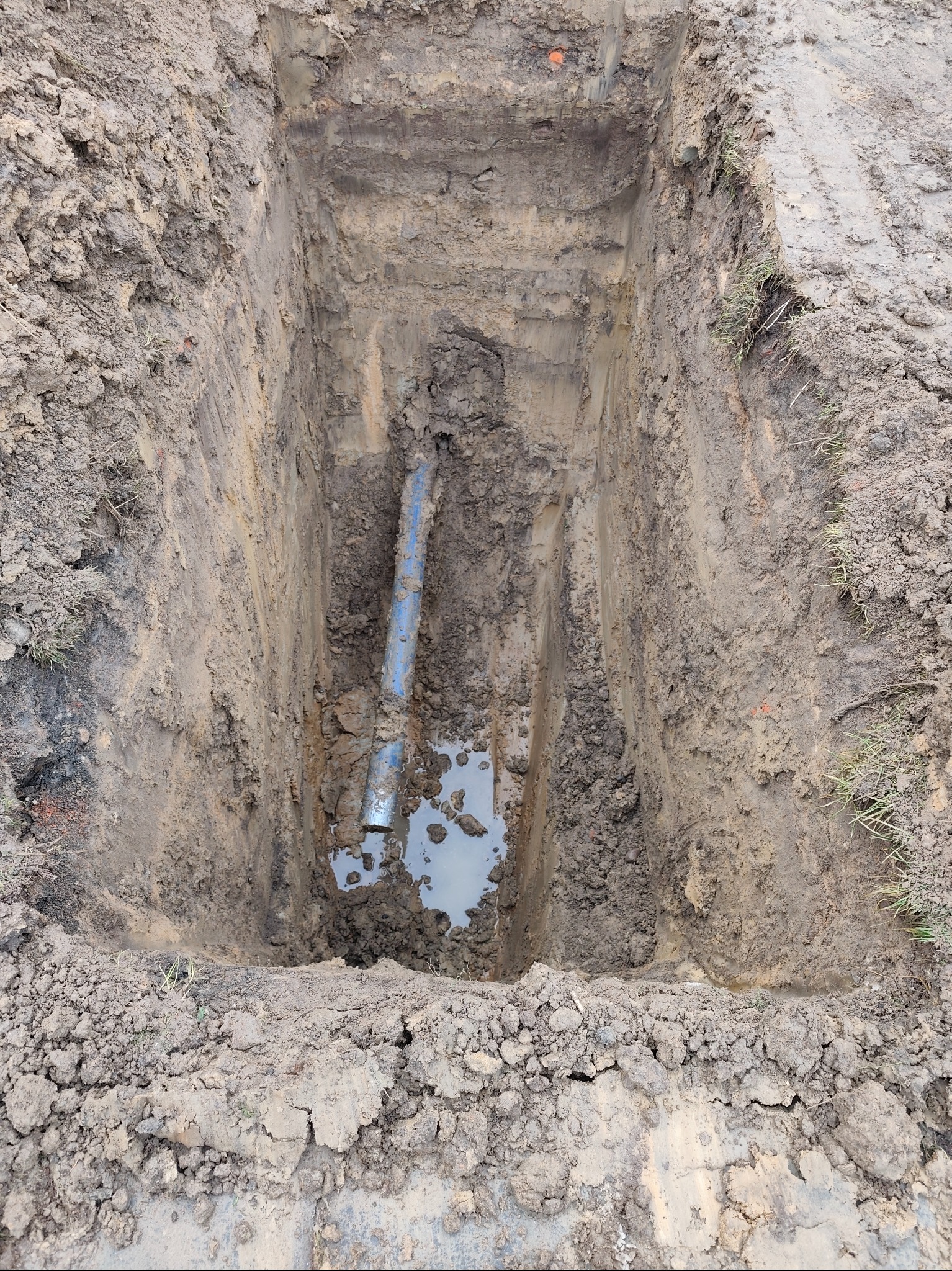 Głęboki wykop w ziemi z widoczną niebieską rurą instalacyjną i wodą na dnie. Ślady opon na powierzchni gruntu.