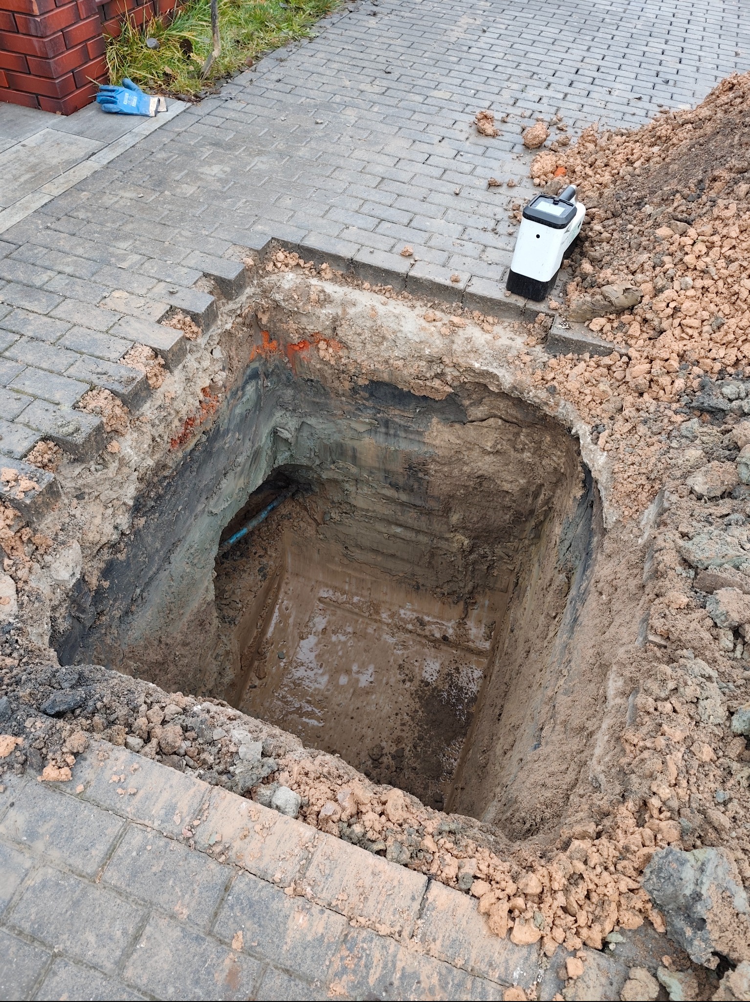 Wykopany dół pod instalację na posesji z kostki brukowej. Widoczna rura w ziemi, urządzenie pomiarowe i rękawice robocze. Prace ziemne w toku.