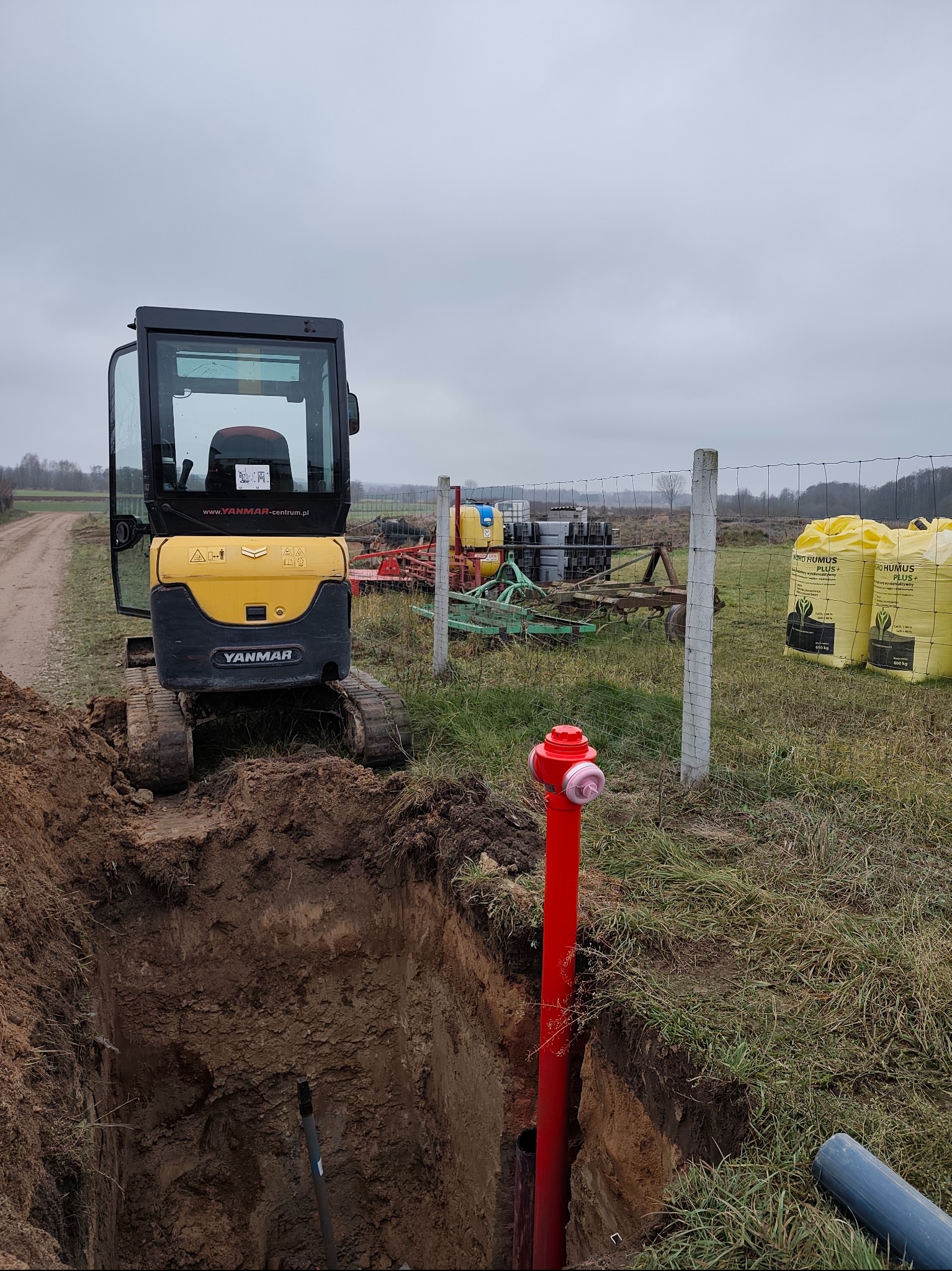 Żółta mini koparka Yanmar przy wykopie z czerwoną rurą i niebieskim przewodem. W tle sprzęt rolniczy i worki z nawozem. Pochmurny dzień na wsi.