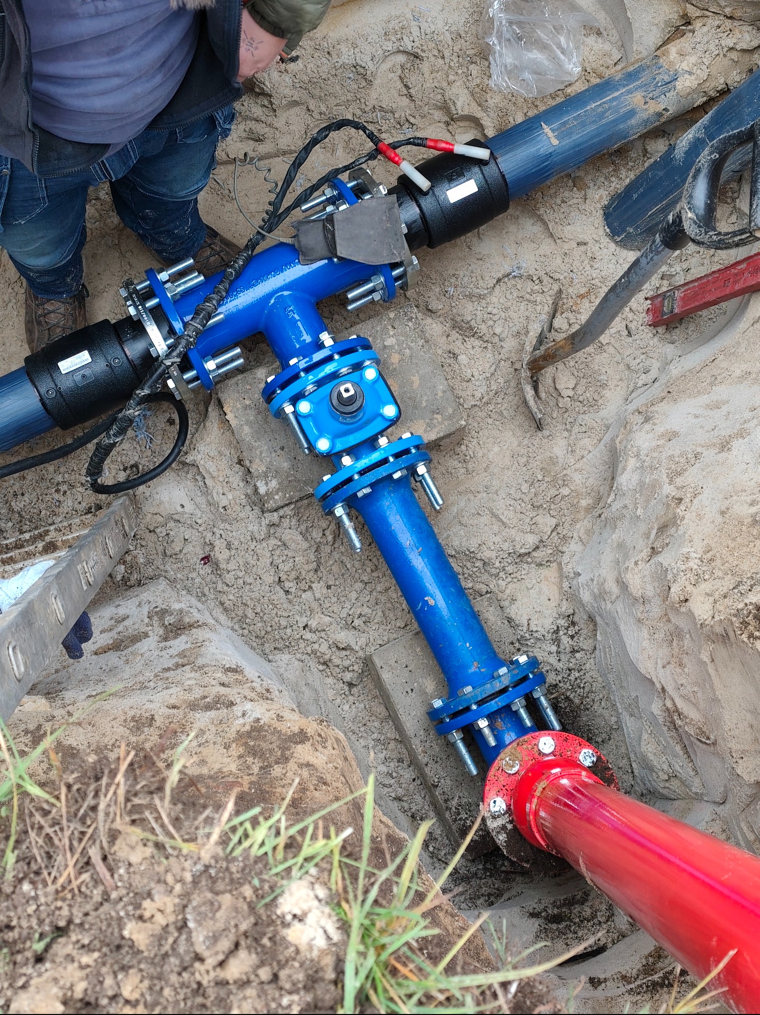 Niebieski trójnik z rurami w wykopie, połączony z czerwoną rurą. Widoczne narzędzia i fragment drabiny. Instalacja wodociągowa w piasku.