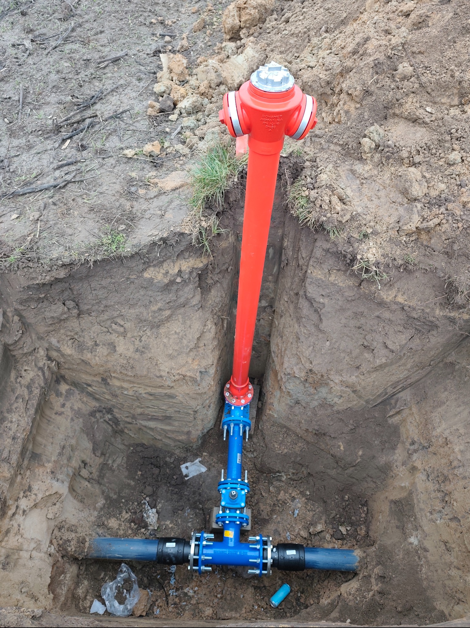 Czerwony hydrant nadziemny połączony z niebieską instalacją w wykopie. Prace instalacyjne w Łodzi. Widok z góry na ziemny wykop.