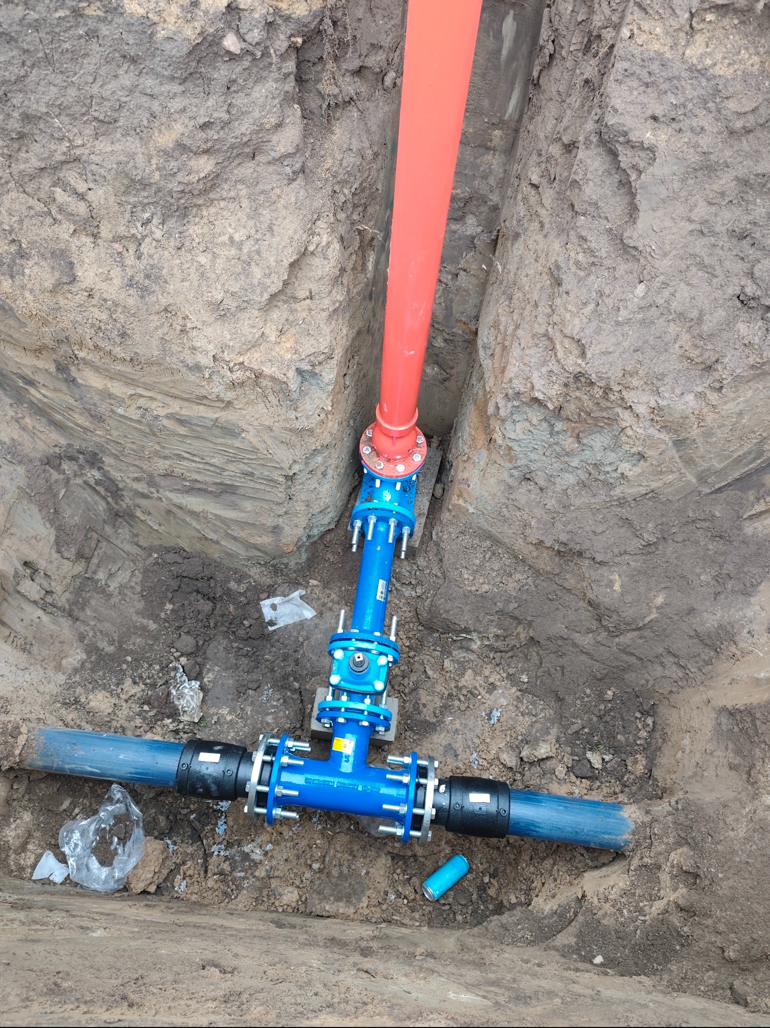 Złącze rur: czerwona pionowa i niebieska pozioma, połączone niebieskim trójnikiem w wykopie ziemnym. Widoczne śruby i uszczelki. Element instalacji podziemnej.