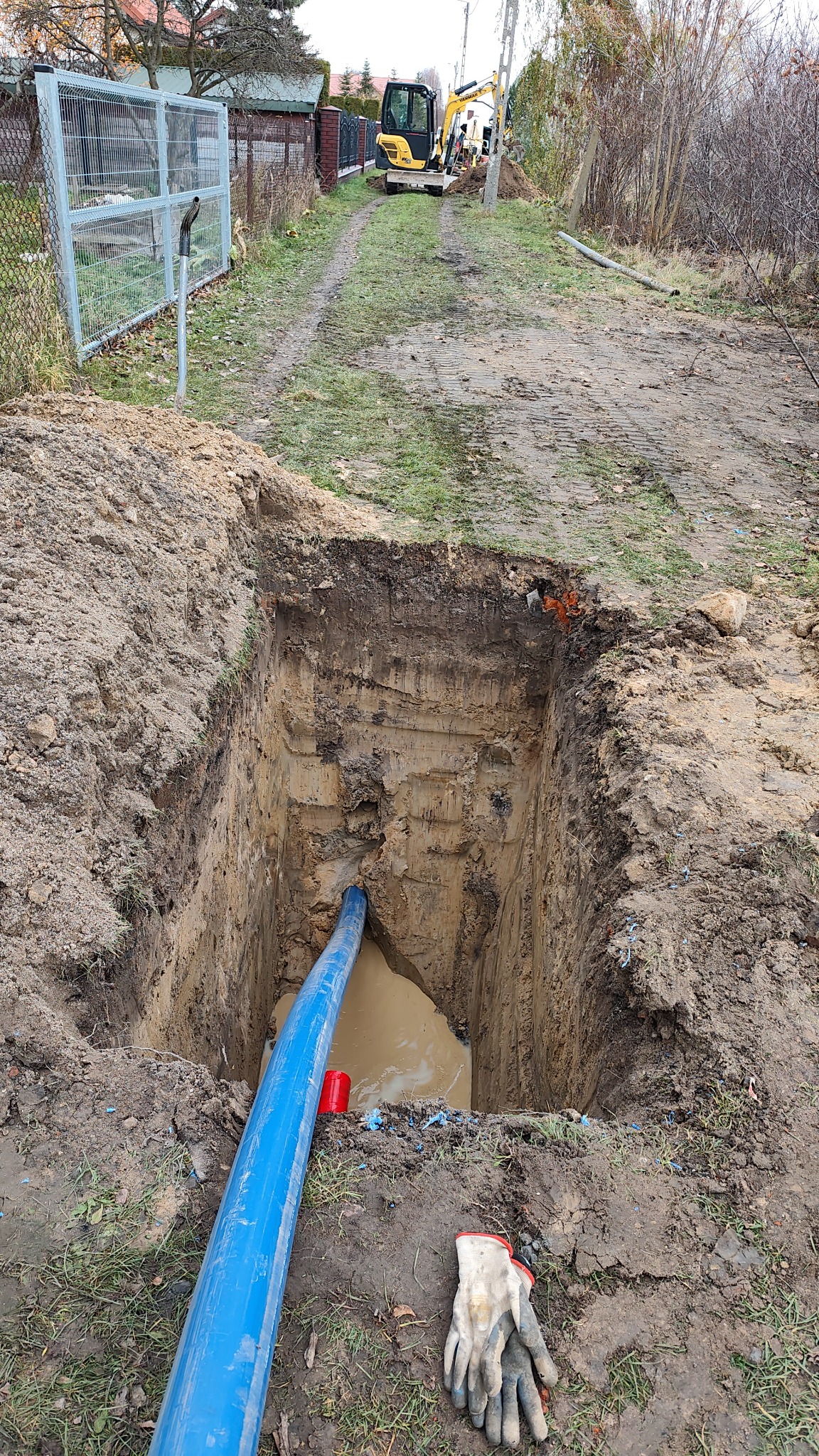 Głęboki wykop z niebieską rurą w środku, prawdopodobnie przyłącze wodne. W tle widoczny mały, żółty koparka i ogrodzenie posesji. Obok wykopu leżą robocze rękawice.