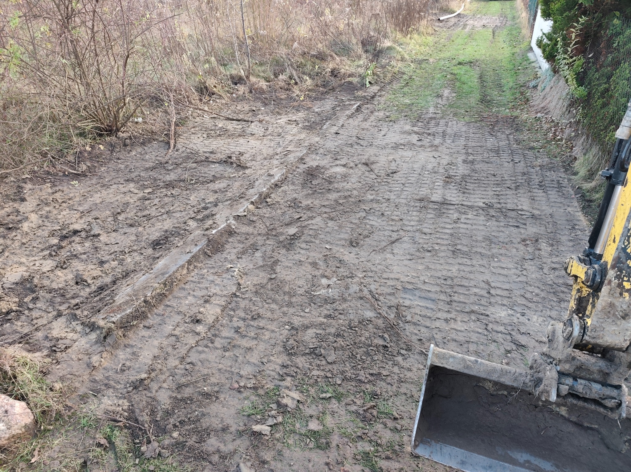 Ślady opon na błotnistej drodze po wykopie, widoczny fragment łyżki koparki. Teren przygotowany pod instalacje.