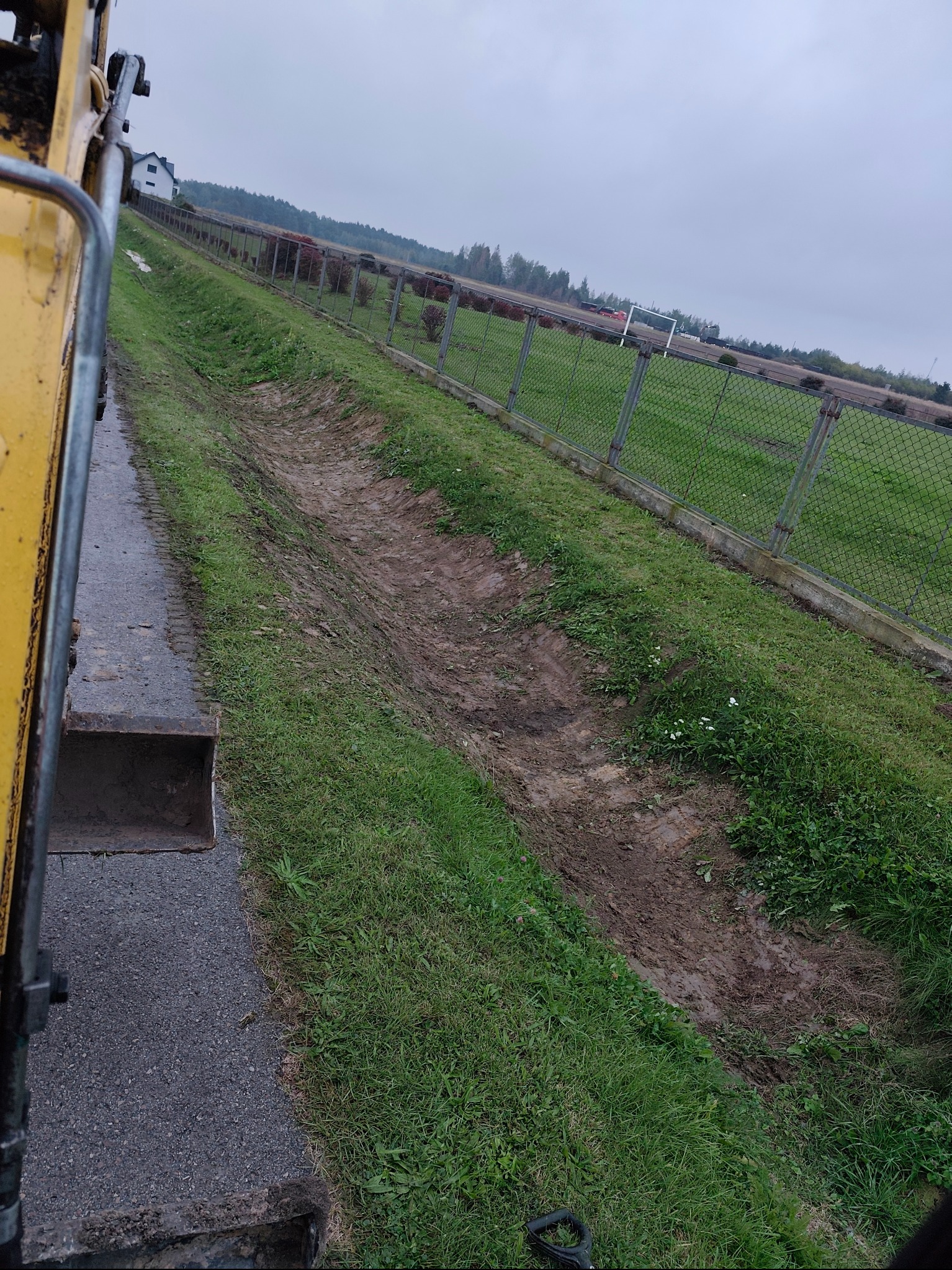 Wykopany rów w ziemi, widok z perspektywy operatora koparki. W tle ogrodzenie z siatki i budynek. Pochmurne niebo.