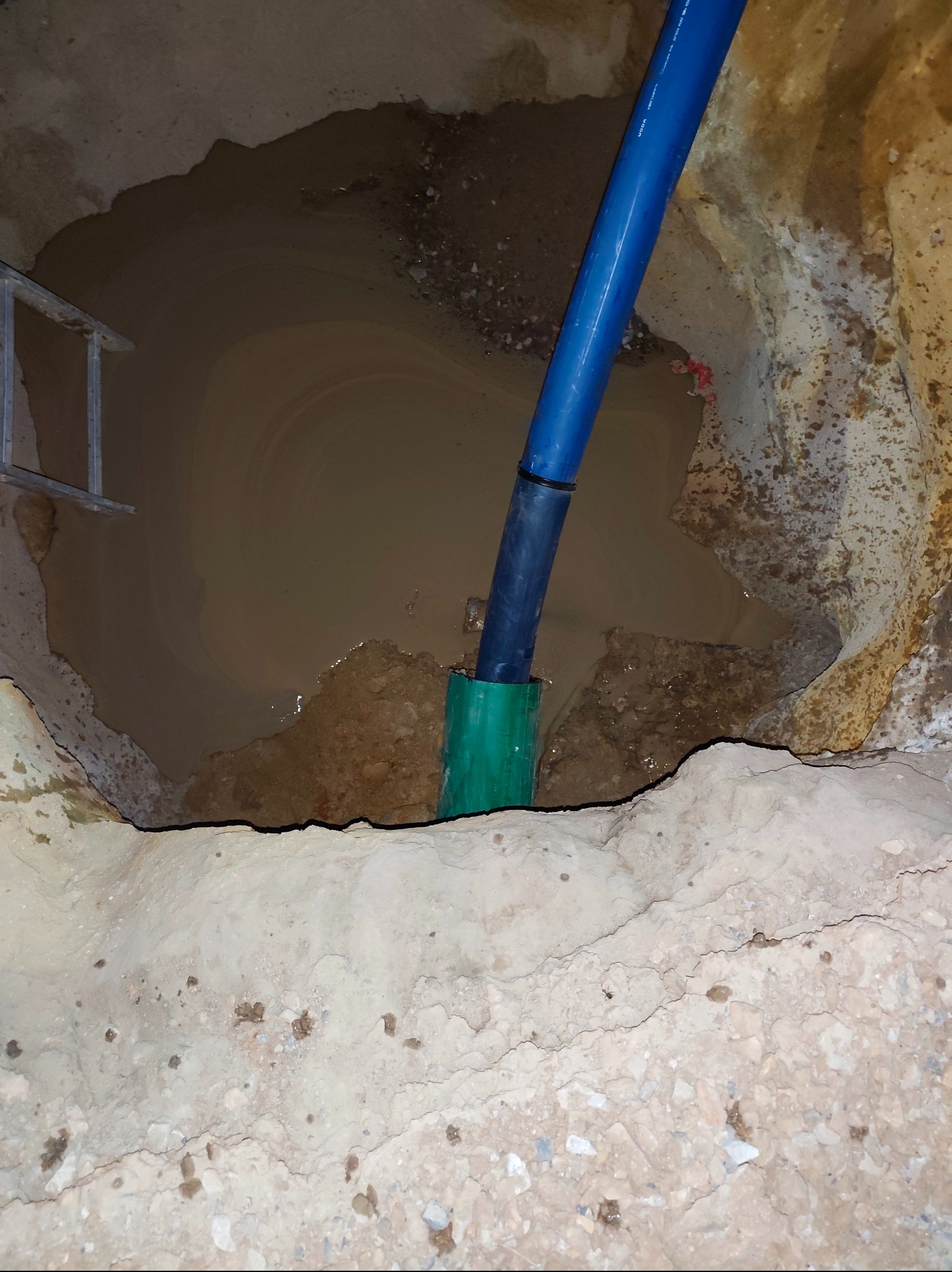 Nietypowe ujęcie: Niebieska rura w zielonej osłonie w wykopie wypełnionym wodą. Widoczna drabina i struktura ziemi. Prace instalacyjne w trudnych warunkach.