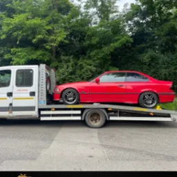 Czerwone BMW E36 na lawecie pomocy drogowej, z logo AHA Auto Holiday Assistance, na tle zieleni. Widok z boku, światło dzienne.