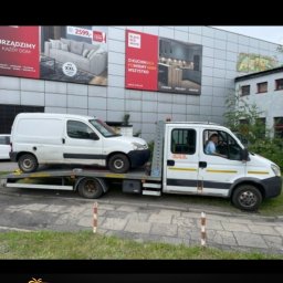 Auto Holiday Assistance Damian Witczak - Biała laweta z białym busem na platformie, reklamy mebli i kuchni w tle. Widoczny kierowca w kabinie. Pomoc drogowa AHA z numerem telefonu.