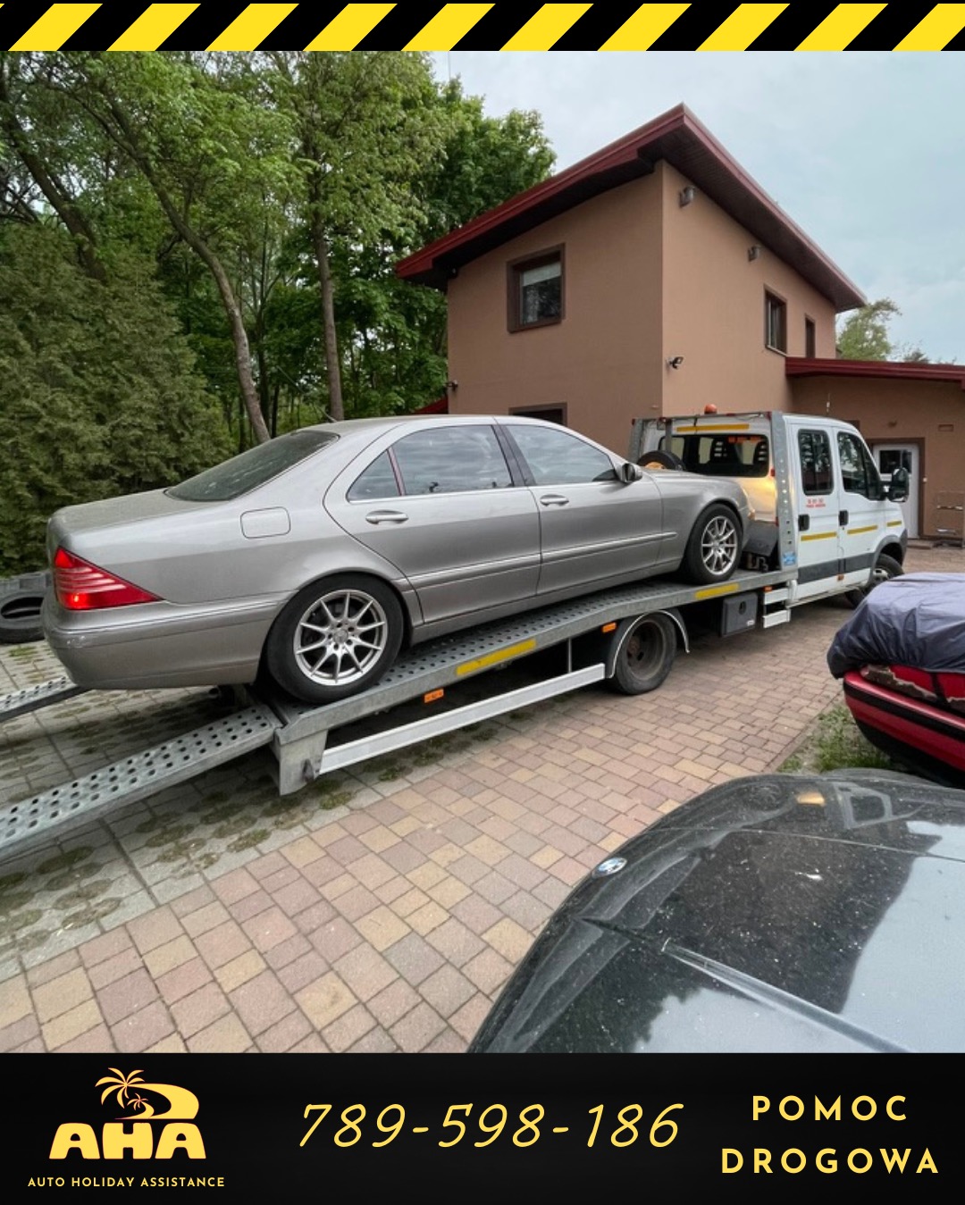 Srebrny samochód osobowy na lawecie w Borowej, z podniesioną platformą. Laweta zaparkowana na tle budynku z czerwonym dachem i zieleni. Widoczne logo firmy.