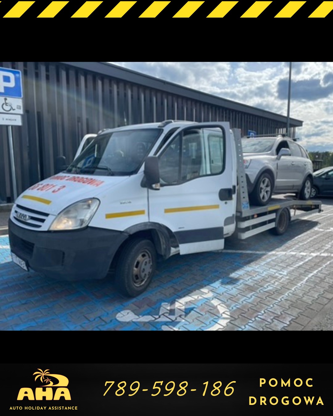 Biała laweta z załadowanym srebrnym SUV-em, zaparkowana na niebieskim parkingu dla niepełnosprawnych, przed budynkiem z szarymi panelami.