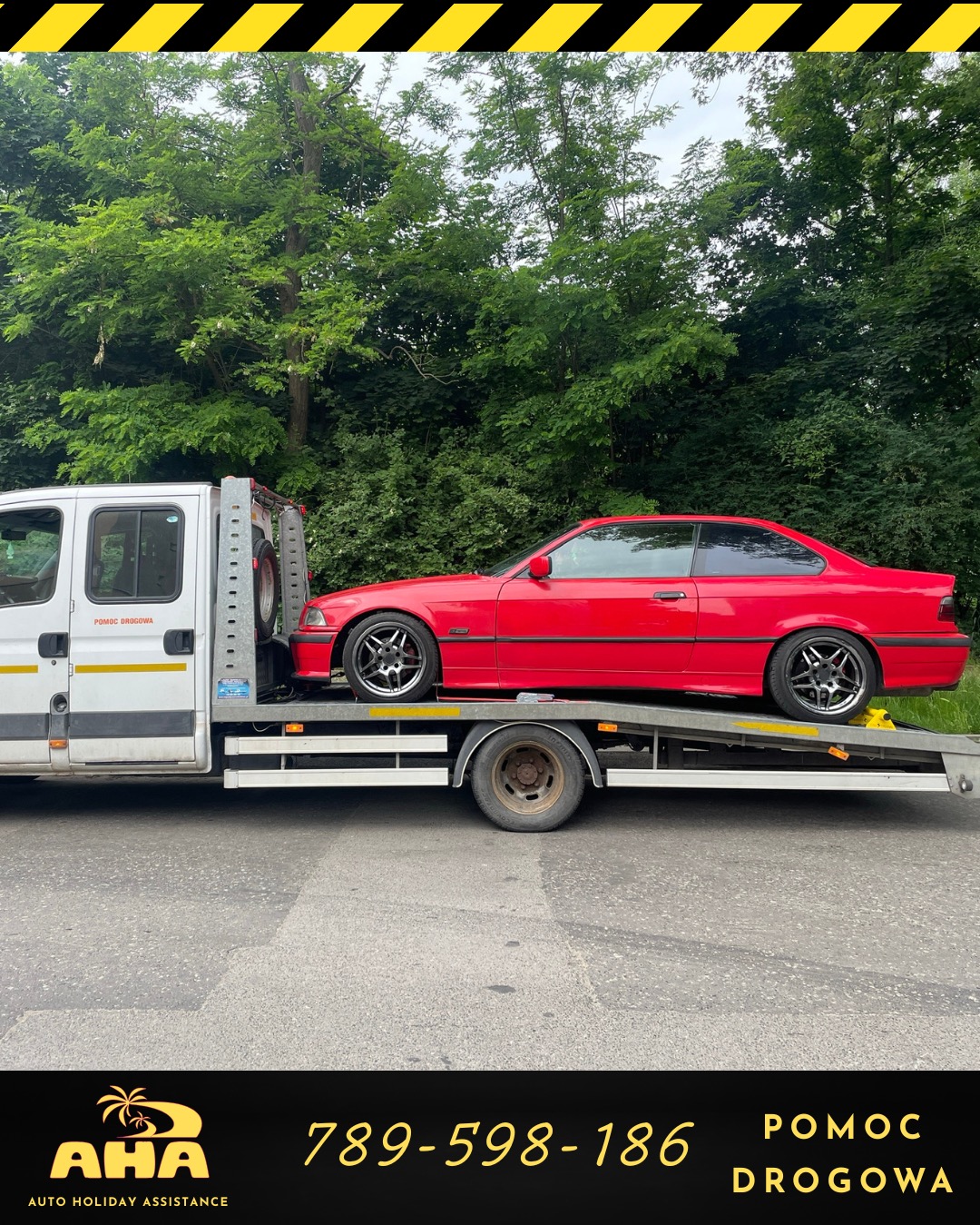 Czerwone BMW E36 na lawecie pomocy drogowej, z logo AHA Auto Holiday Assistance, na tle zieleni. Widok z boku, światło dzienne.