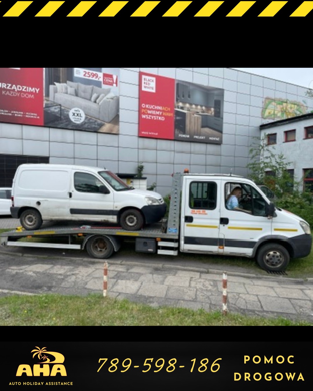 Biała laweta z białym busem na platformie, reklamy mebli i kuchni w tle. Widoczny kierowca w kabinie. Pomoc drogowa AHA z numerem telefonu.