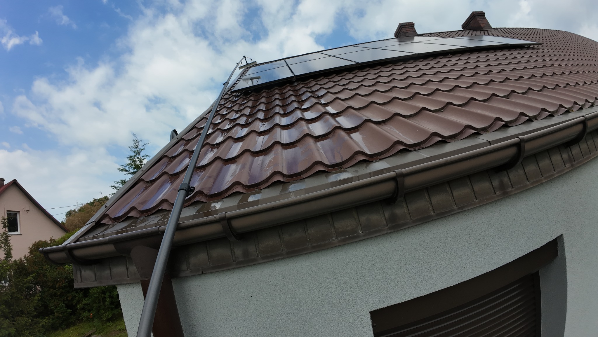 Czerwono-brązowy dach z panelami słonecznymi, czyszczony lancą teleskopową, z widoczną rynną i fragmentem elewacji na tle błękitnego nieba.