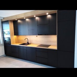 Usługi stolarskie Marcin Araźny - Nowoczesna kuchnia z czarnymi frontami szafek i drewnianym blatem, oświetlona punktowo. Widoczny zlew i płyta indukcyjna. Minimalistyczny design.