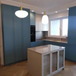 Niebieska zabudowa kuchenna z wyspą na parkiecie w jodełkę. Nowoczesne lampy wiszące. Jasne blaty i białe szafki wiszące. Elegancki minimalizm.