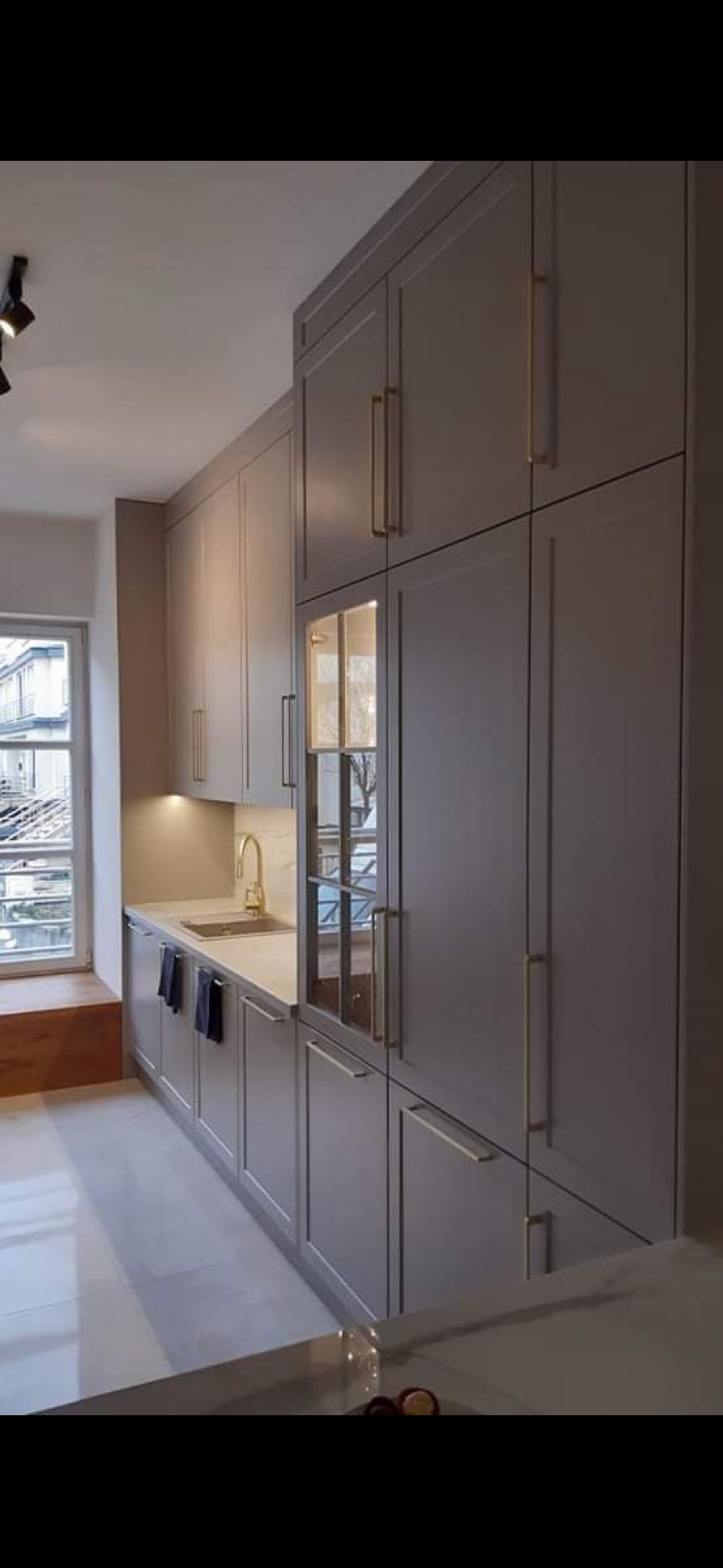 Szara zabudowa kuchenna z złotymi uchwytami i szklaną witryną, widok z dołu, Ciechanowiec. Nowoczesna kuchnia na wymiar z oknem i drewnianym siedziskiem.