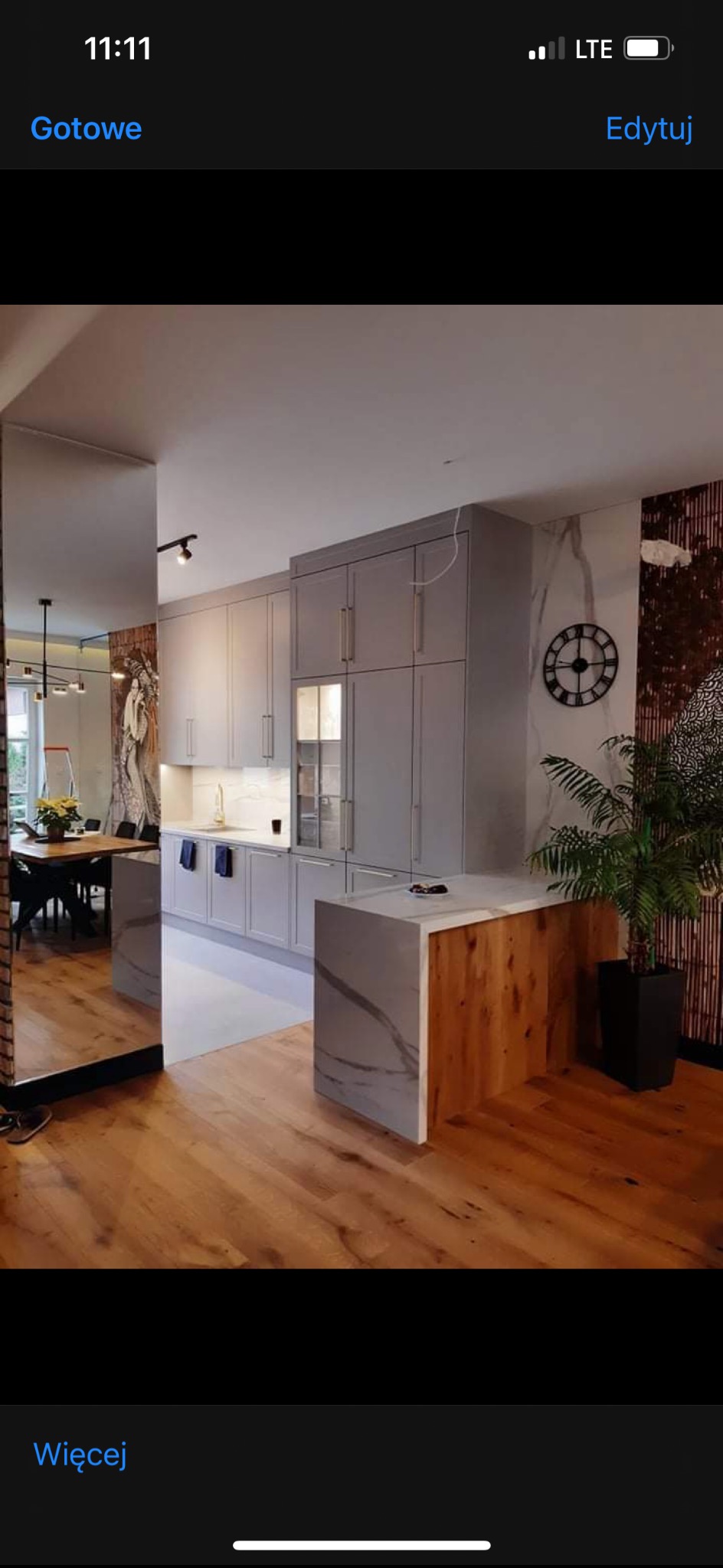 Nowoczesna kuchnia w odcieniach szarości z drewnianymi akcentami i marmurowym blatem. Widoczny stół z krzesłami i roślina doniczkowa. Całość dopełnia dekoracja ścienna.