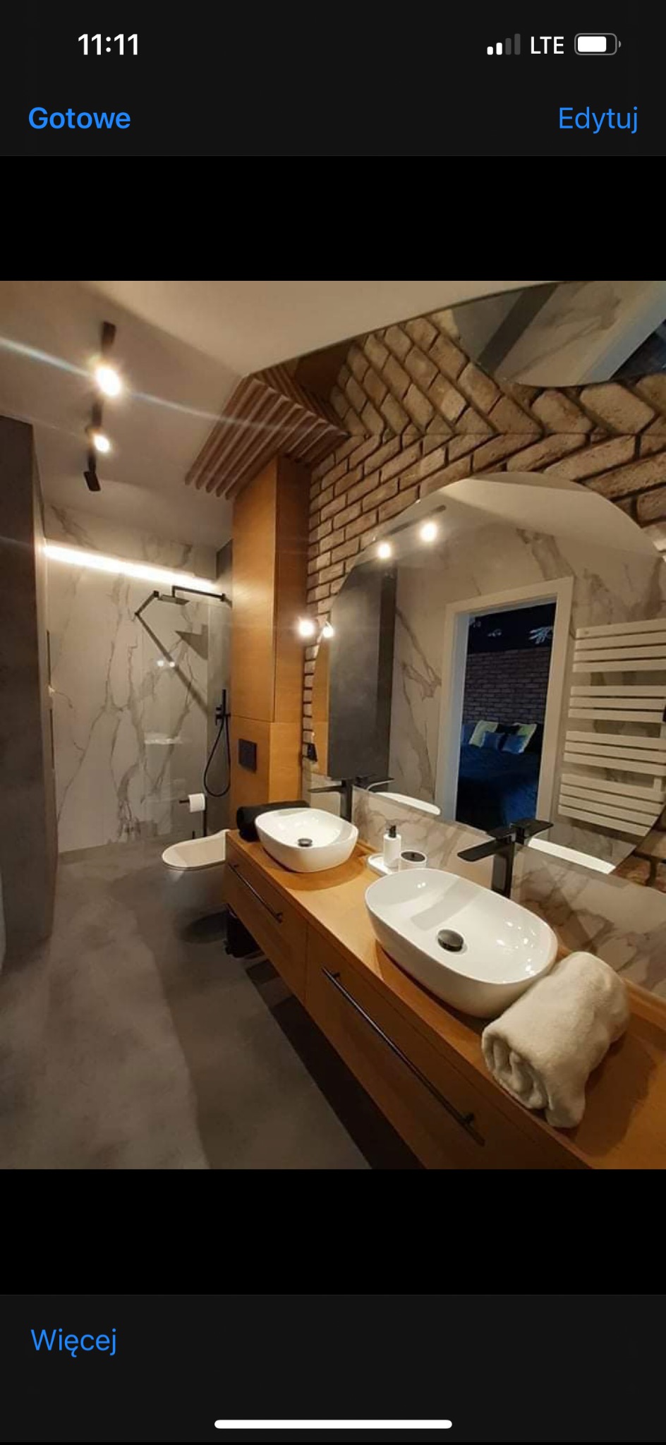 Nowoczesna łazienka z drewnianą szafką pod umywalki, lustrem w ozdobnej ramie i elementami cegły na ścianie. Widoczny prysznic i toaleta w minimalistycznym stylu.