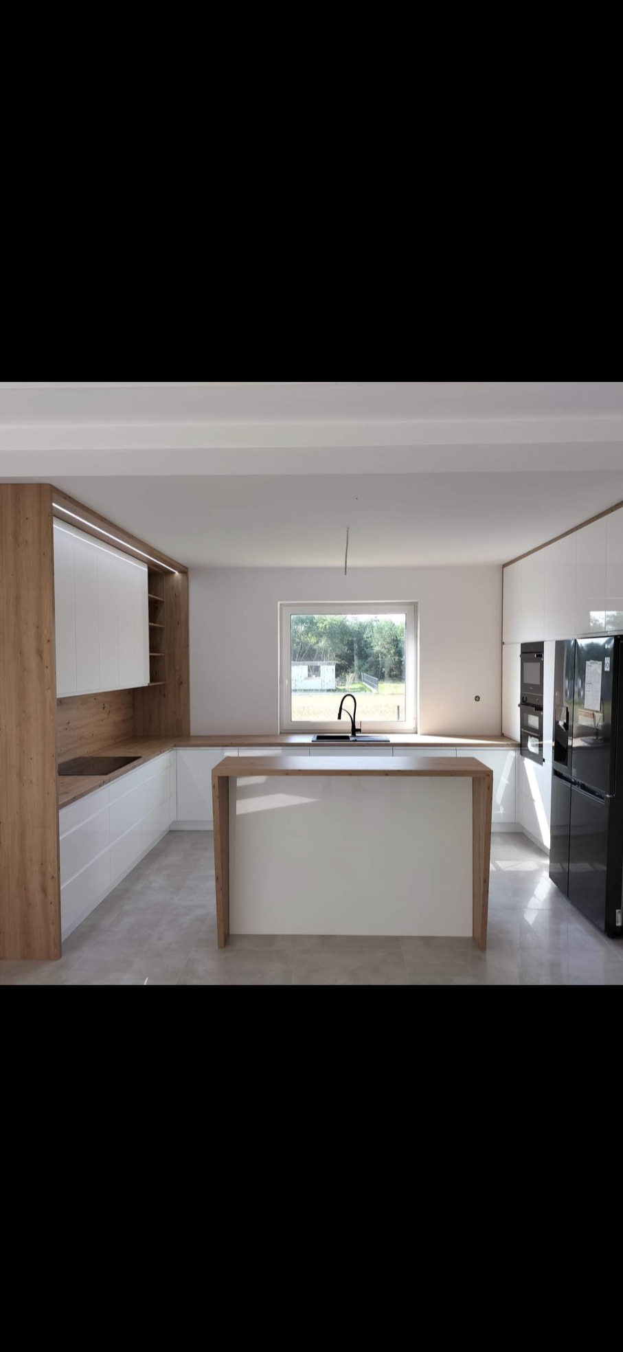 Nowoczesna kuchnia z wyspą, białe fronty i drewniane akcenty, czarna armatura, minimalistyczne oświetlenie, duże okno z widokiem na zieleń.