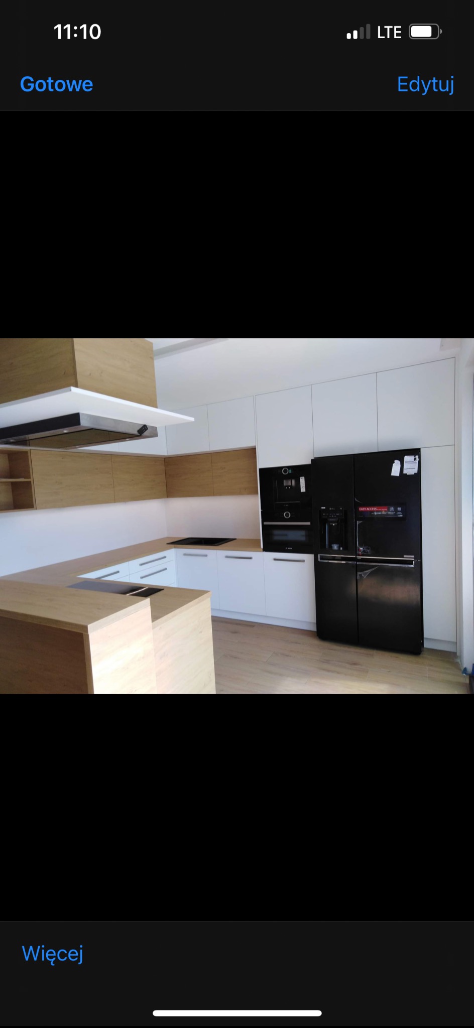 Jasna kuchnia z drewnianymi akcentami i czarnym sprzętem AGD, widok z perspektywy blatu, minimalistyczny design, nowoczesne oświetlenie.