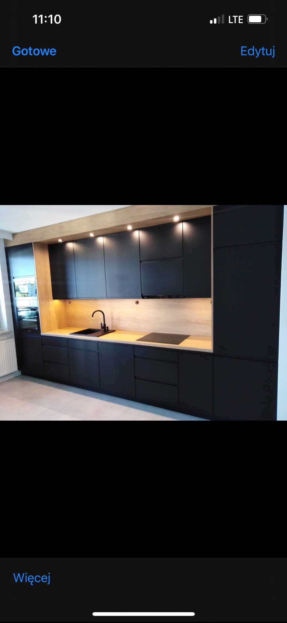 Nowoczesna kuchnia z czarnymi frontami szafek i drewnianym blatem, oświetlona punktowo. Widoczny zlew i płyta indukcyjna. Minimalistyczny design.