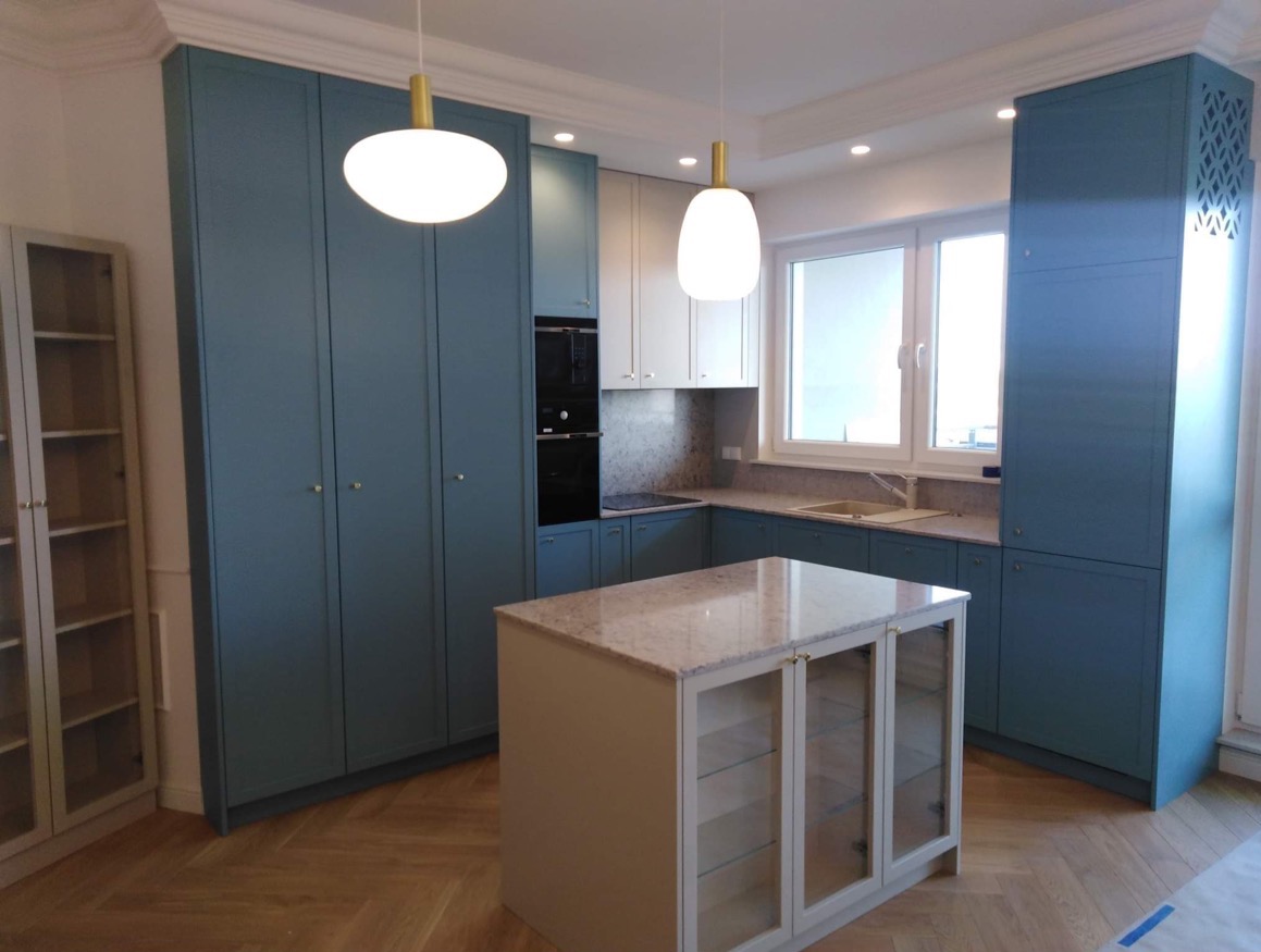 Niebieska zabudowa kuchenna z wyspą na parkiecie w jodełkę. Nowoczesne lampy wiszące. Jasne blaty i białe szafki wiszące. Elegancki minimalizm.