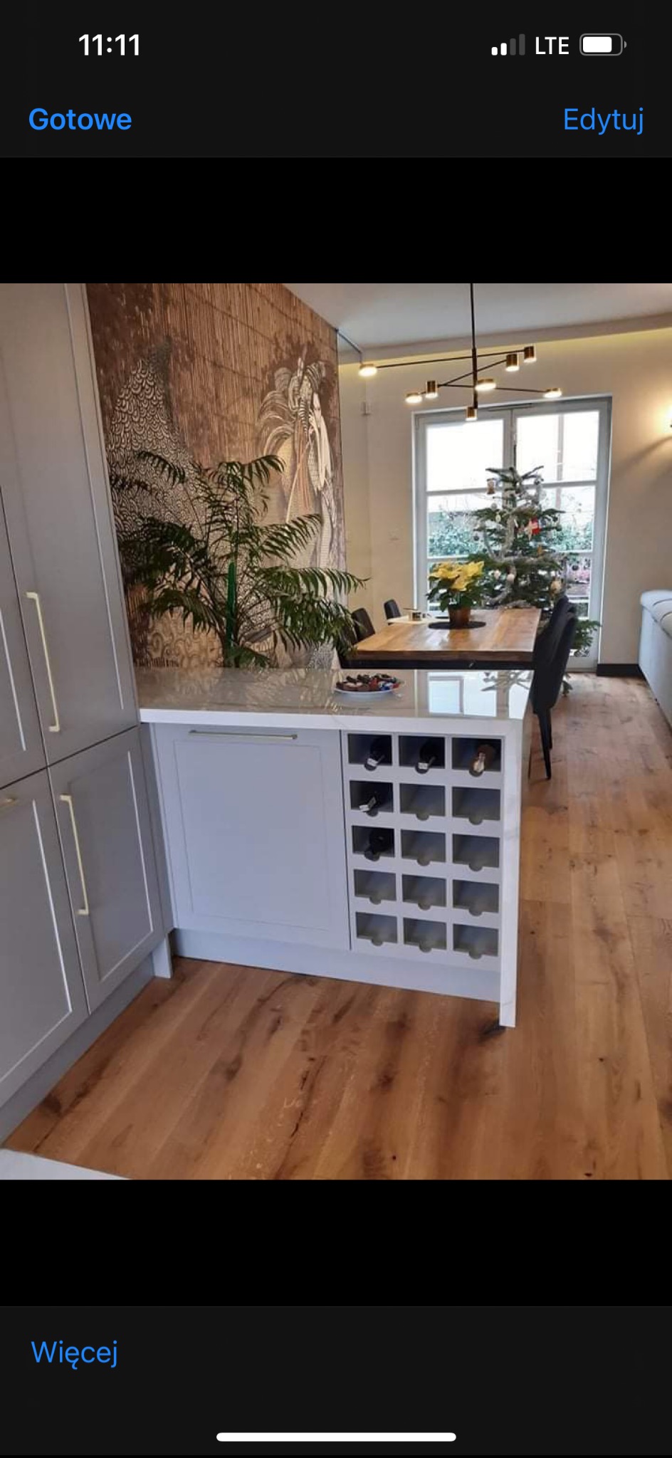 Jasnoszara wyspa kuchenna z półkami na wino, drewniana podłoga i tapeta z motywem leśnym w tle. Widoczny stół z krzesłami i choinka.