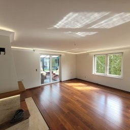 MatHouse - Jasne, przestronne wnętrze po remoncie: parkiet z egzotycznego drewna, białe ściany z listwami LED pod sufitem, duże okno z widokiem na zieleń, kominek z podświetleniem.