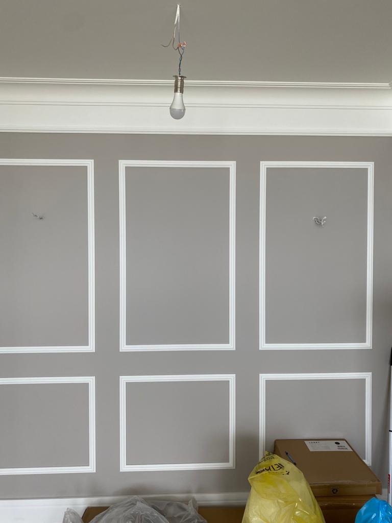 Ściana w szarym kolorze z białymi ozdobnymi listwami tworzącymi prostokąty. W górnej części ściany biały gzyms. Wisząca żarówka na kablu. Częściowo widoczne materiały budowlane.