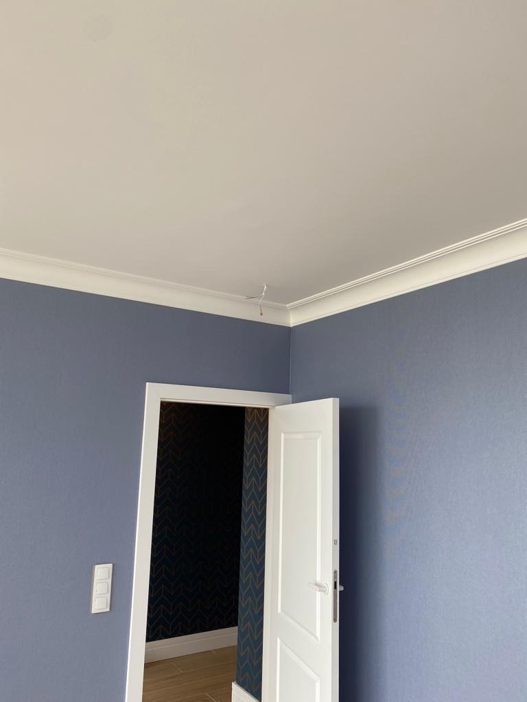 Niebieskie ściany z białą sztukaterią i listwami przypodłogowymi, otwarte białe drzwi z widoczną tapetą wewnątrz pokoju, włącznik światła na ścianie.