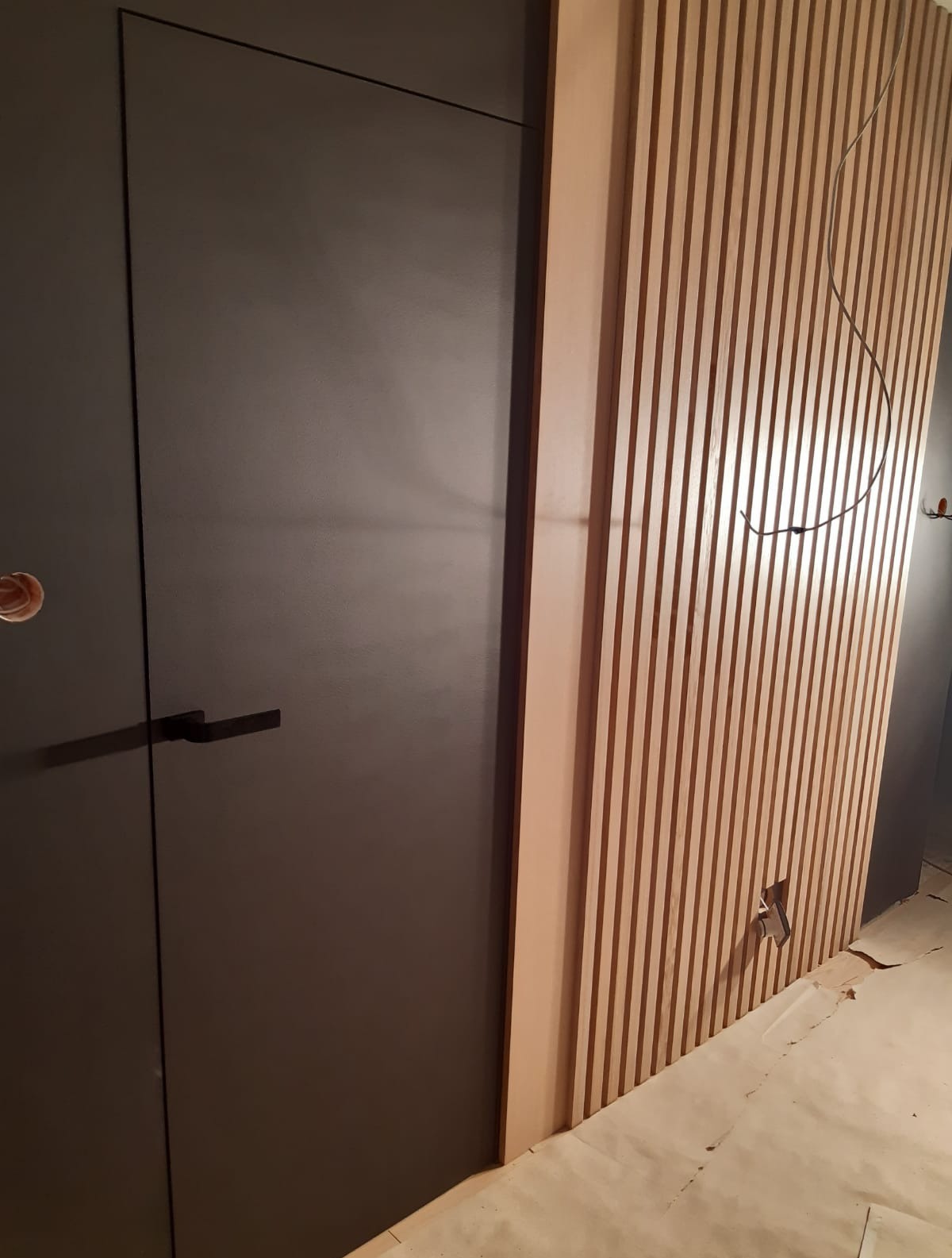 Nowoczesne drzwi w kolorze antracytu z czarną klamką, sąsiadujące z dekoracyjną ścianą z drewnianych lameli. Minimalistyczny design z widocznymi elementami instalacji.