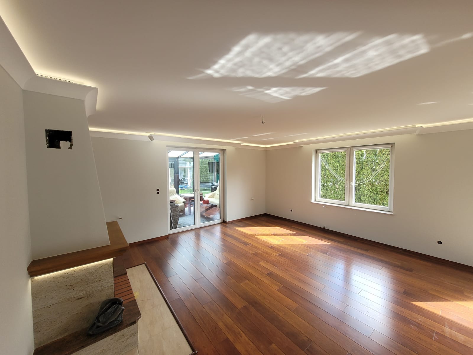 Jasne, przestronne wnętrze po remoncie: parkiet z egzotycznego drewna, białe ściany z listwami LED pod sufitem, duże okno z widokiem na zieleń, kominek z podświetleniem.