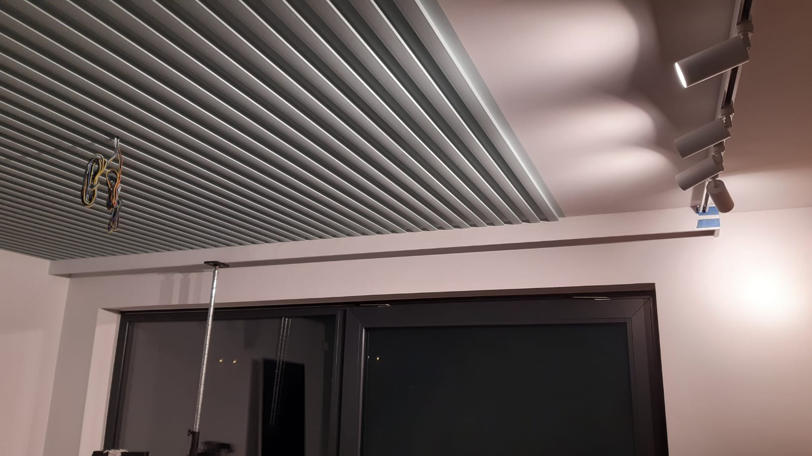 Nowoczesny sufit z metalowych paneli i szynoprzewodem z oświetleniem punktowym. Widoczne wiszące kable i okno.