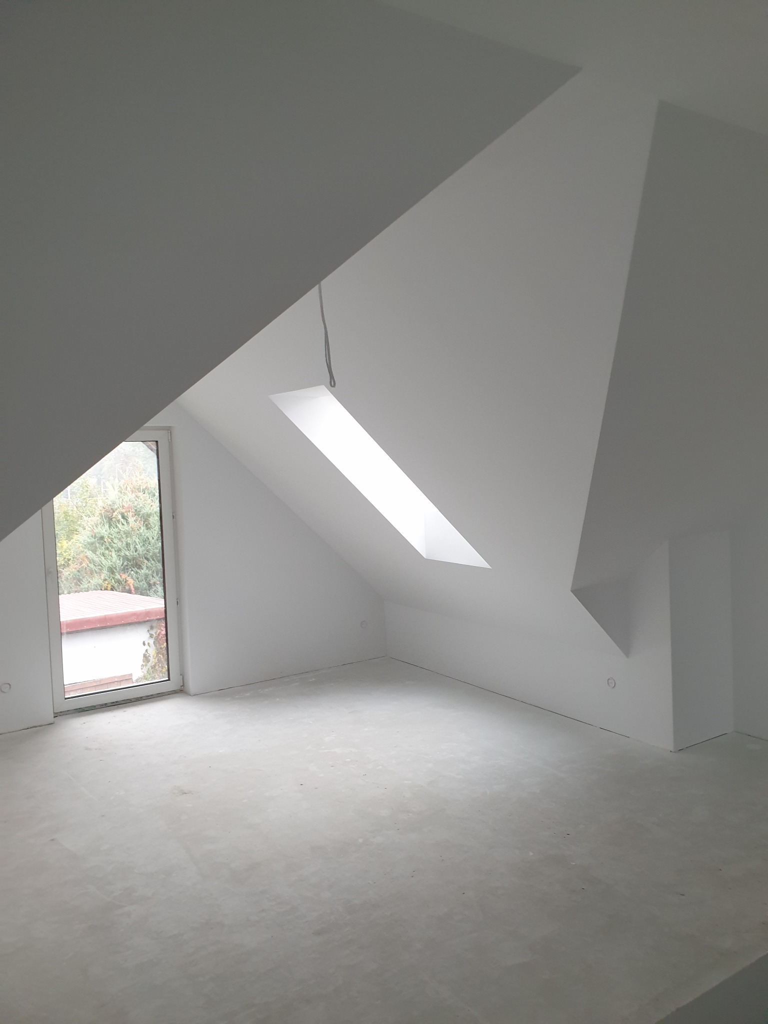 Białe poddasze ze świetlikiem i oknem. Ściany i sufit pomalowane na biało, surowa wylewka na podłodze. Widok na zieleń za oknem.