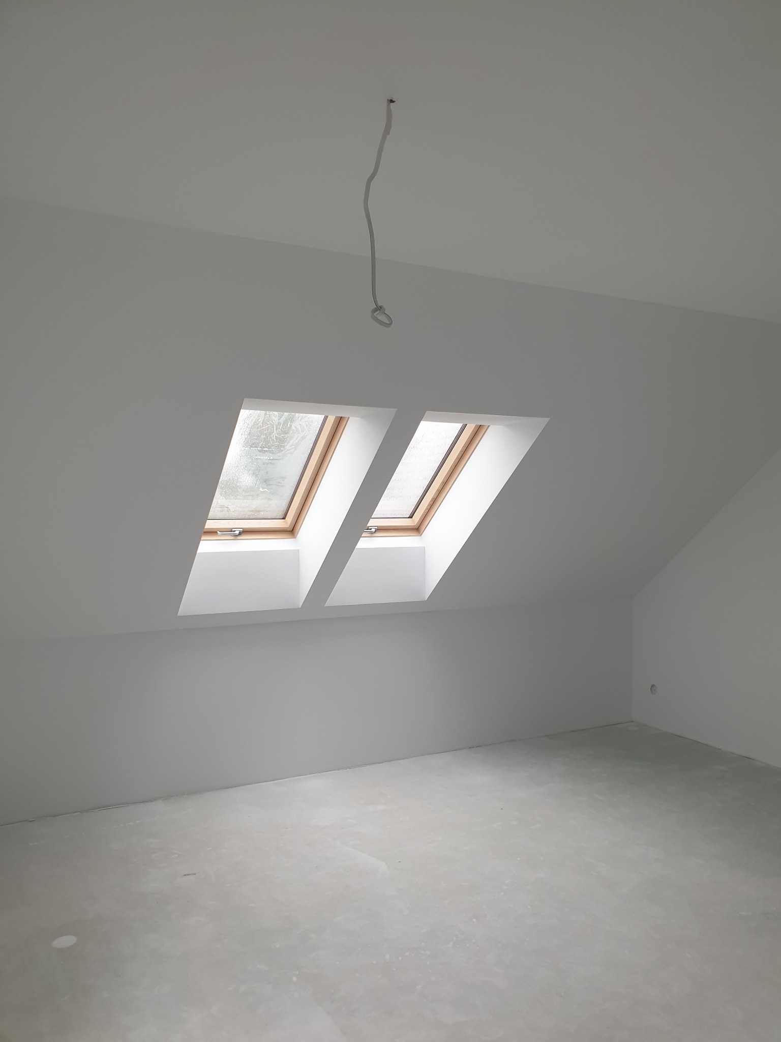 Poddasze po remoncie: białe ściany, dwa okna dachowe z drewnianą ramą, surowa podłoga, wiszący przewód elektryczny. Minimalistyczne wykończenie.